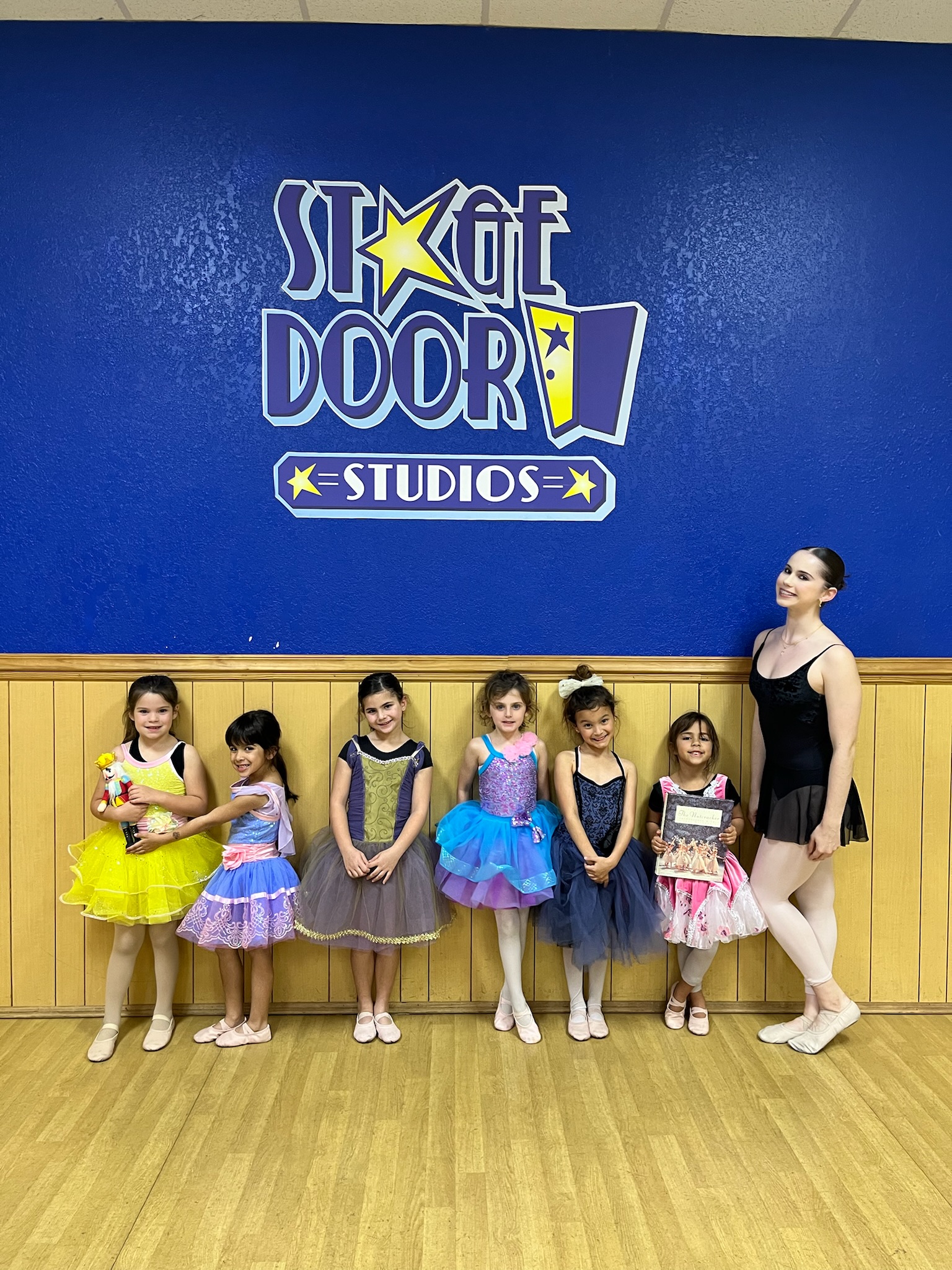Young Stage Door Studios ballet students in colorful tutus posing with their teacher in front of the studio logo